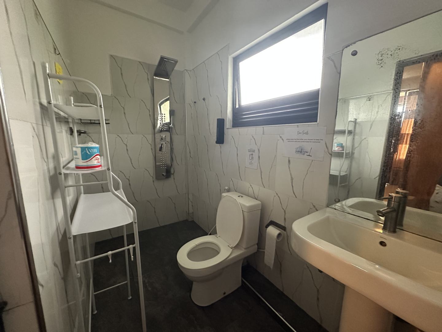 A wet room in marble-veined tile with a rain-shower panel, a basin, a mirror and a shelf stacked with paper.