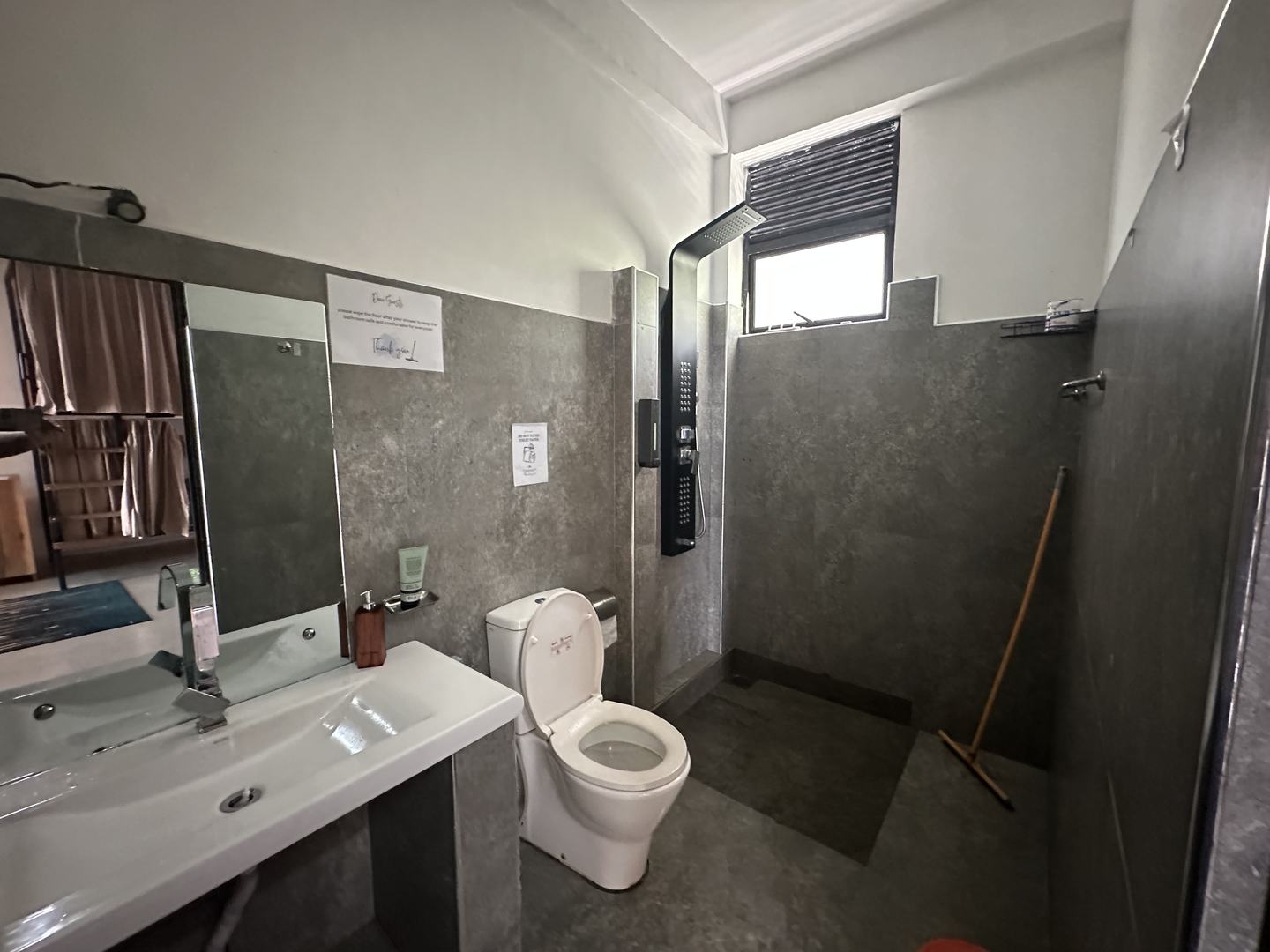 A grey-tiled wet room with a black rain-shower panel, a basin, a toilet and a window above the shower.