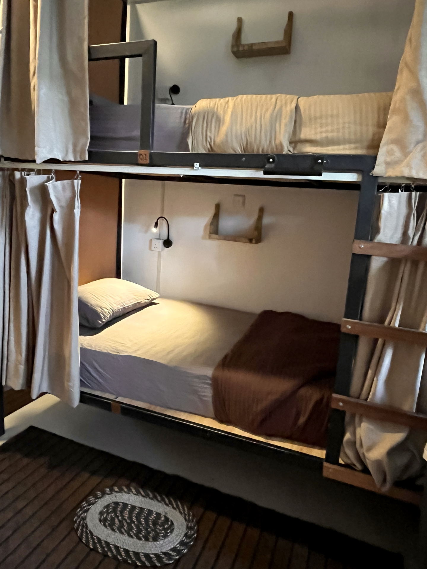 A lower bunk with its curtain drawn back: a reading lamp, a power socket and a small wooden shelf at the head of the bed.