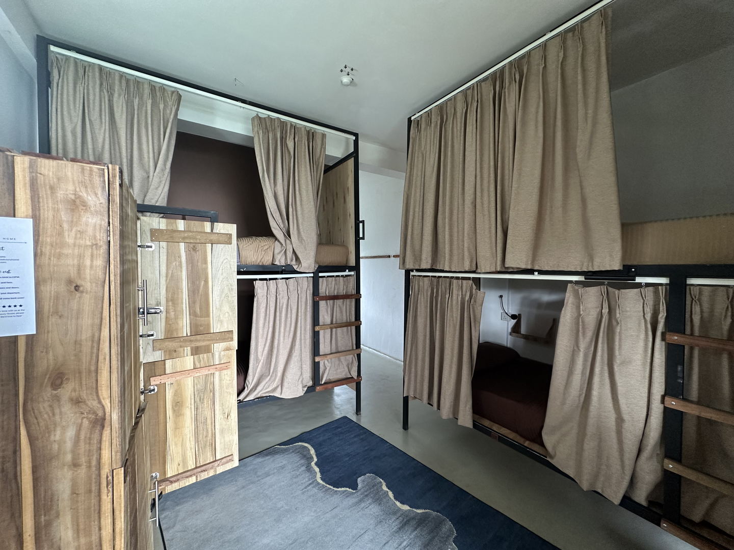 A dormitory seen from the doorway: curtained bunks on both sides, a timber wardrobe with a laminated notice taped to it, and a rug on the floor.