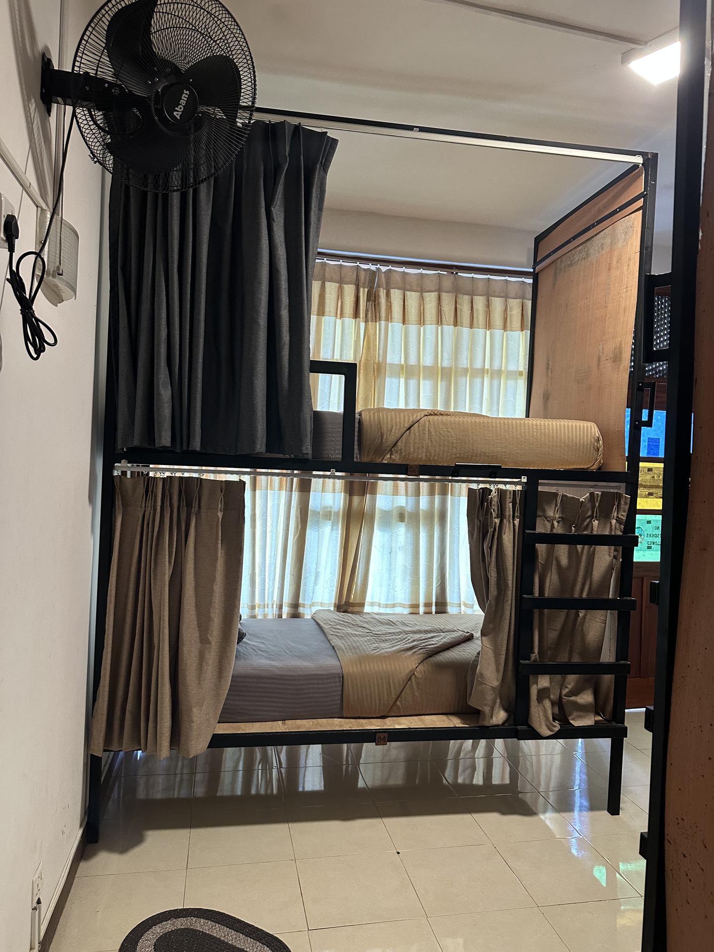 A bunk in a four-bed mixed dorm with the curtains drawn back, a wall fan above it and a curtained window behind.