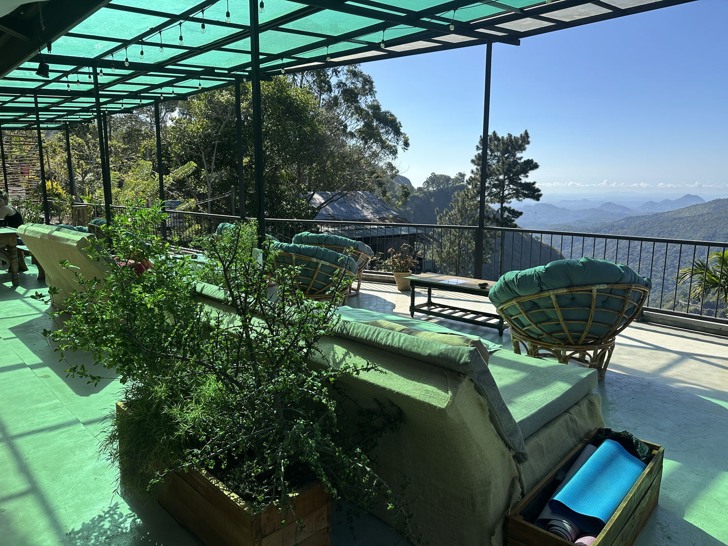 Built-in benches with teal cushions and a round rattan chair under the green roof, a planter of herbs in front and the valley past the railing.