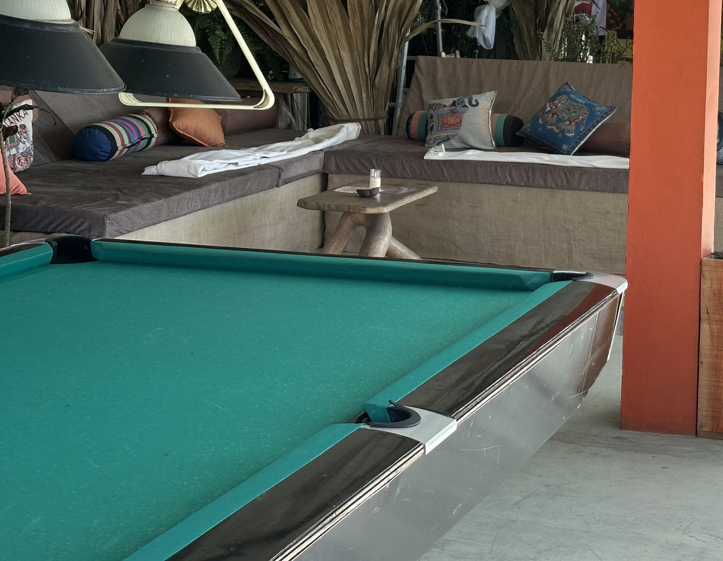 A pool table under hanging metal lamps, with a cushioned banquette behind it, dried-palm columns and a red-painted pillar.