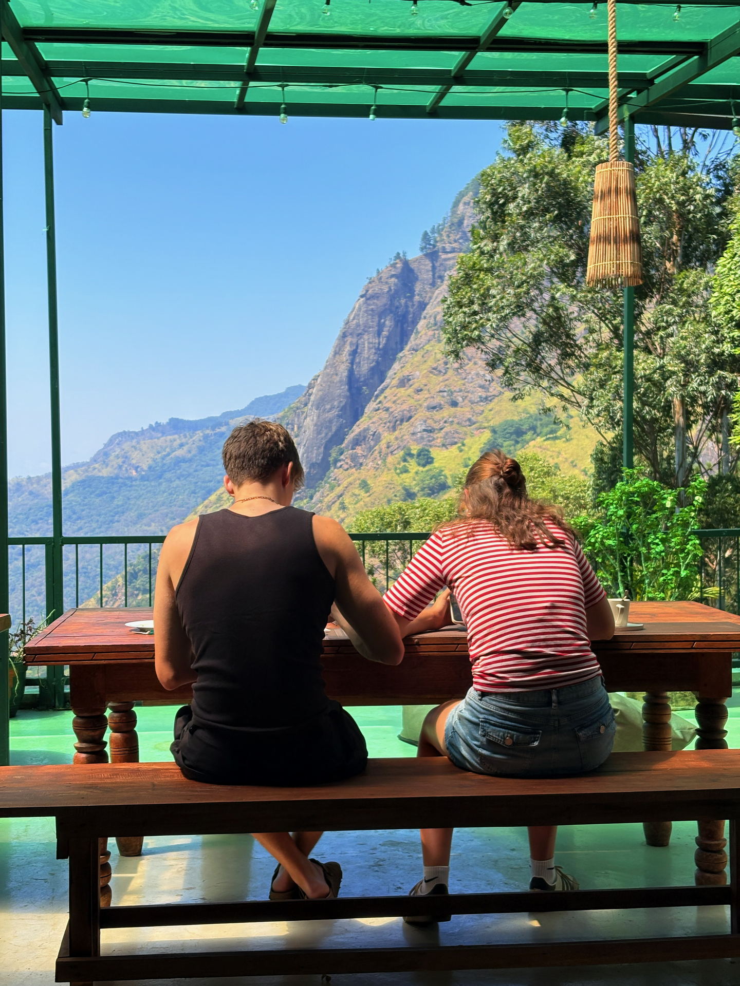 Two people sitting at a long timber table on the hostel’s rooftop terrace, seen from behind, facing a mountain ridge across a valley.