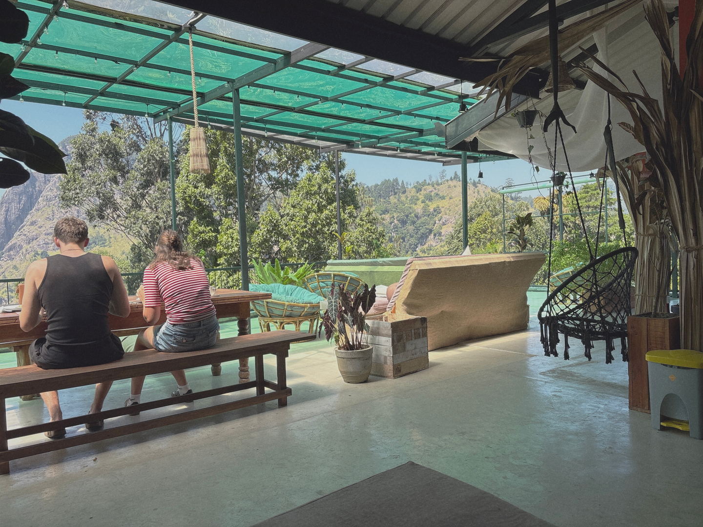 The terrace under its green roof: two people seen from behind at a long timber table, benches, a hanging chair and the valley past the railing.