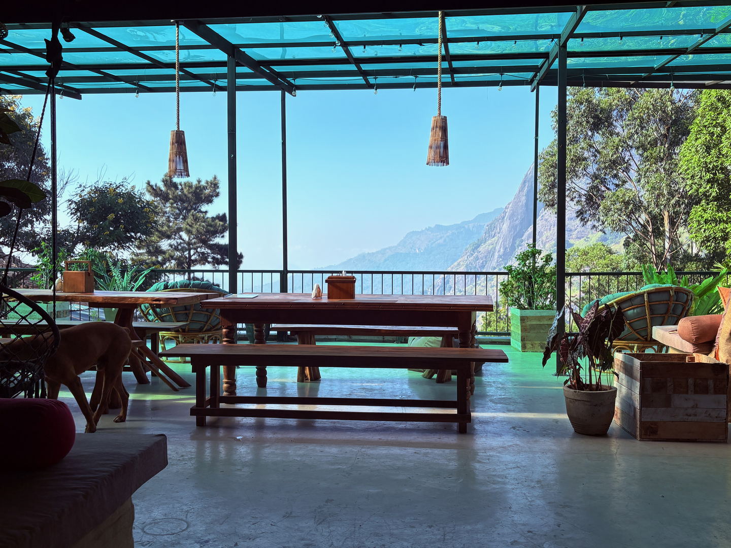 The open rooftop terrace: long timber tables and benches under a translucent green roof on a black steel frame, with no wall on the valley side.